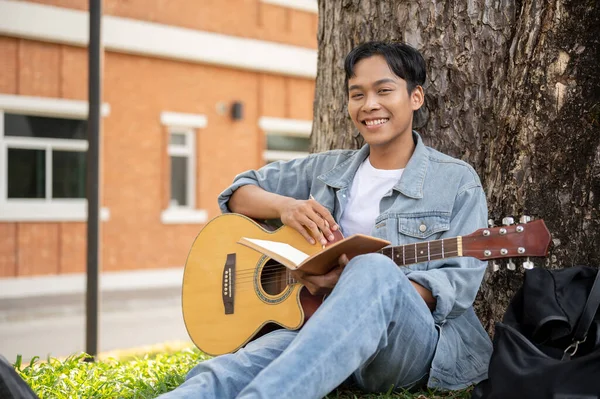 Genç pozitif Asyalı erkek, akustik gitarıyla bir parkta ağacın altında dinleniyor ve gitar çalıyor. Boş zaman ve hobi konsepti