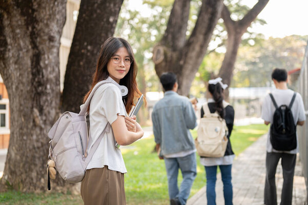 A positive, cute Asian female student in casual clothes with her laptop computer is walking on a footpath on her campus, going to school in the morning. Back to school concept