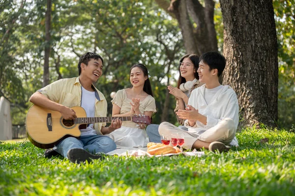 A group of cheerful young Asian friends are enjoying a picnic in a green park together, singing and playing guitar, eating yummy foods, and having a fun summer day together. Friendship concept