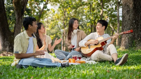 A group of cheerful young Asian friends are enjoying a picnic in a green park together, singing and playing guitar, eating yummy foods, and having a fun summer day together. Friendship concept