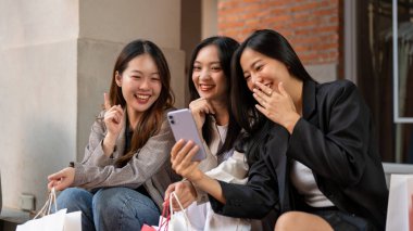 Bir grup neşeli, eğlenceli Asyalı bayan arkadaş şehirde merdivenlerde birlikte otururken akıllı telefondan eğlenceli bir şeyler izliyorlar. Alışveriş günü, seyahat, şehir hayatı.
