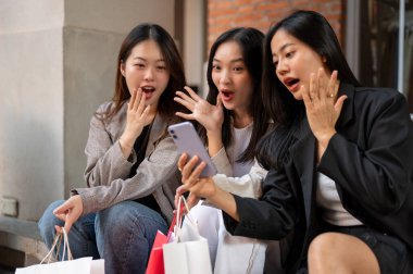 Bir grup heyecanlı ve şaşırmış Asyalı bayan arkadaş şehirde merdivenlerde otururken beklenmedik online indirimle şok olmuş, şaşırtıcı yüzleri olan bir akıllı telefon ekranına bakıyorlar..
