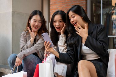 Bir grup heyecanlı ve şaşırmış Asyalı bayan arkadaş şehirde merdivenlerde otururken beklenmedik online indirimle şok olmuş, şaşırtıcı yüzleri olan bir akıllı telefon ekranına bakıyorlar..