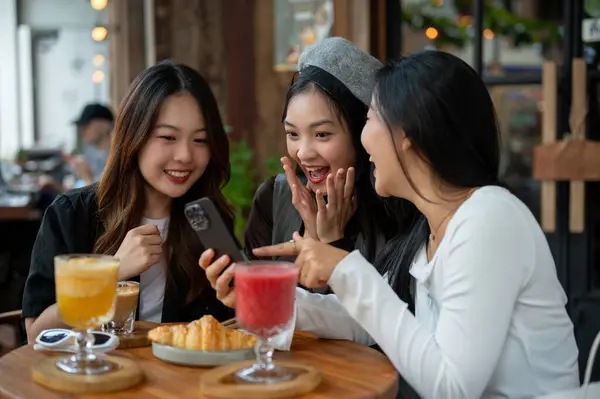 Group of attractive Asian female friends are surprised with some news online on a smartphone and enjoying talking in a cafe or restaurant, hanging out on the weekend.