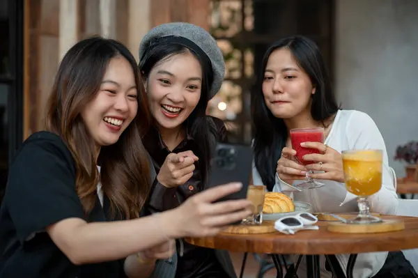 Group of cheerful and attractive Asian female friends are enjoying hanging out at a cafe or restaurant in the city together, chit chat, watching something interesting on a phone together.