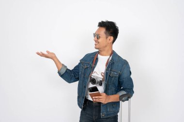 Kot ceketli yakışıklı, mutlu Asyalı bir adam bagajıyla izole edilmiş beyaz bir arka planda dururken avucunu açıyor. seyahat önerisi, tavsiye, sunum