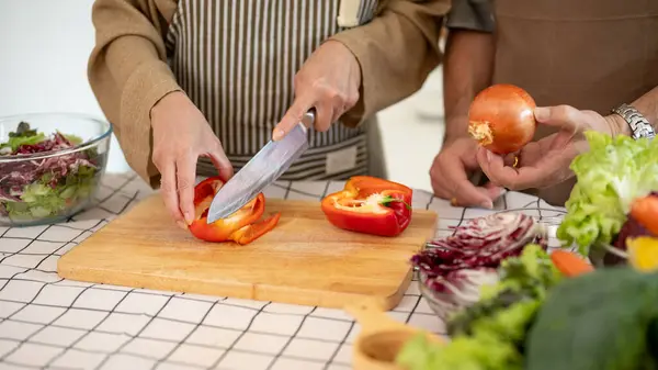 Close-up image of a happy, lovely Asian wife is chopping a sweet pepper while her husband is helping her, enjoy cooking in the kitchen together. home cooking, domestic life, happy quality time