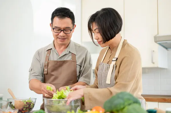 Happy adult Asian couple, husband and wife, are making a salad bowl in the kitchen together, preparing healthy food for their breakfast. home cooking, domestic life, people and food concepts