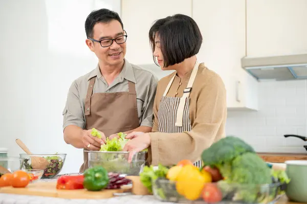 Happy adult Asian couple, husband and wife, are making a salad bowl in the kitchen together, preparing healthy food for their breakfast. home cooking, domestic life, people and food concepts