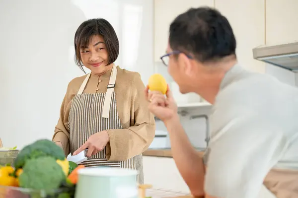 A happy Asian husband is enjoying talking with his wife while she is cooking, making breakfast in the kitchen. family bonding, home cooking, domestic life, adult couple