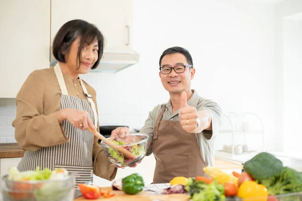 A happy adult Asian husband is showing his thumb up to the camera while enjoying cooking in the kitchen together. family bonding, home cooking, domestic life, adult couple