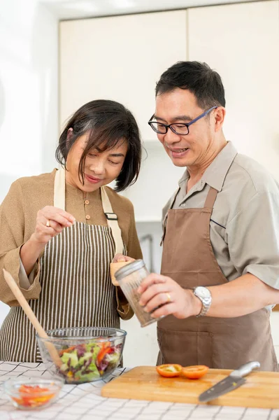 Happy Asian couples, husband and wife, are cooking a healthy salad bowl in the kitchen together, adding seasoning, and enjoying the weekend at home. Home cooking, life partners, domestic life