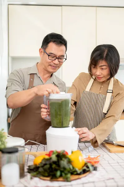 Happy adult Asian couples are enjoying making healthy green smoothie in the kitchen together, using a smoothie blender. Home cooking, family bonding, domestic life