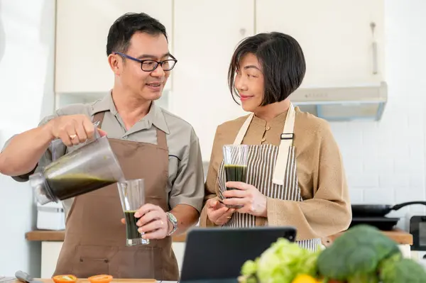 A happy Asian wife and husband are enjoying healthy green smoothie in the kitchen together. Adult couples, domestic life, healthy lifestyle