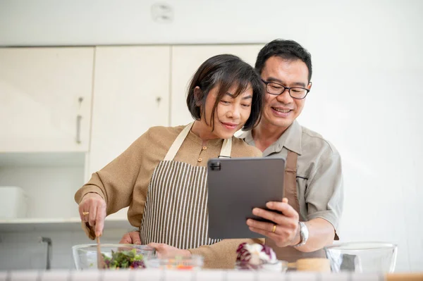 Lovely, happy adult Asian couples are looking at an online recipe on a digital tablet and cooking in the kitchen together. family bonding, home cooking, domestic life, adult couples