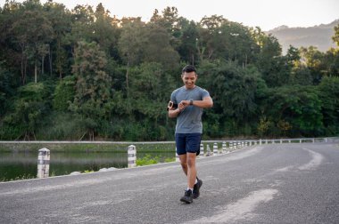 Spor giyimli mutlu ve formda bir Asyalı, parkta koşarken kalp atışlarını kontrol ediyor ve kilometrelerce koşuyor. Sabah sporu, açık hava egzersizi, koşu, maraton.