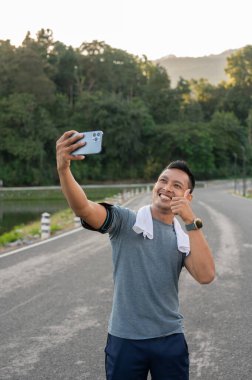 Spor kıyafetleri içinde mutlu, yakışıklı bir Asyalı sabah koşusu sırasında parmağını gösterip selfie çekiyor ya da videosunu kaydediyor..