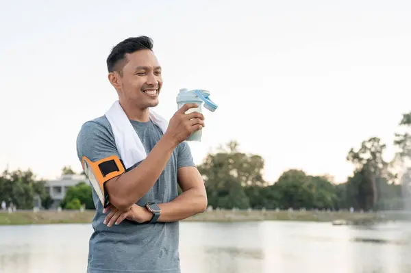 A happy, strong and fit Asian man in sportswear is drinking a protein shake after a long run outdoors, having a sport drink supplement for muscle gain.