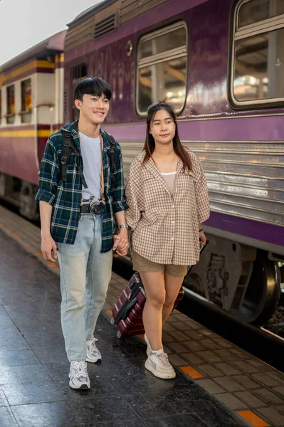 Young happy Asian couple travelers are commuting by train together, walking on a platform at a railway station, and catching the train. People and transportation concepts