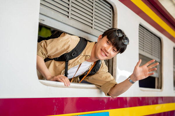 A happy, excited young Asian man traveler backpacker is on a train, smiling and waving his hand, saying goodbye to someone, and traveling somewhere by a train. People and transportation concepts