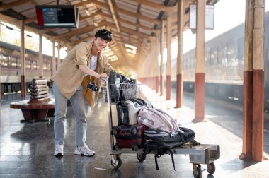 Heyecanlı ve neşeli genç bir Asyalı gezgin elinde bir arabayla platformda tren istasyonunda trenini bekliyor. İnsanlar ve ulaşım kavramları