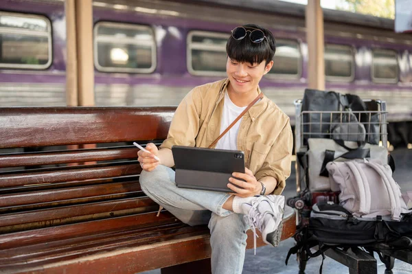 A happy, handsome Asian man is using his digital tablet on a bench while waiting for his train at a railway station. Lifestyle, commuter, public transportation