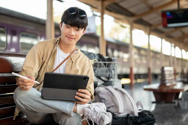 A happy, handsome Asian man is using his digital tablet on a bench while waiting for his train at a railway station. Lifestyle, commuter, public transportation