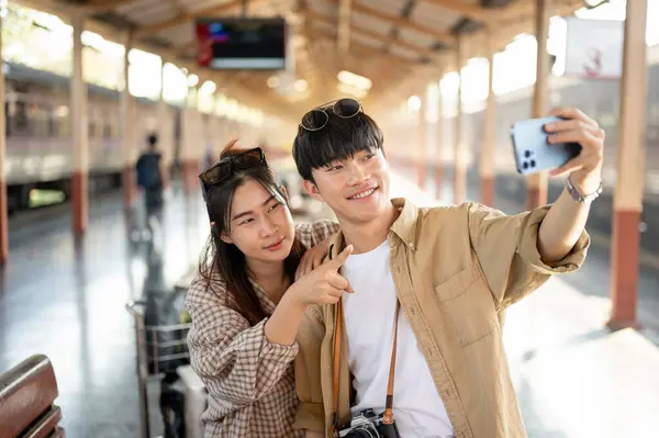 Cute and happy young Asian couple taking selfies or recording a video with a smartphone while waiting for the train at a railway station together, excited for their summer trip. Lifestyle concept