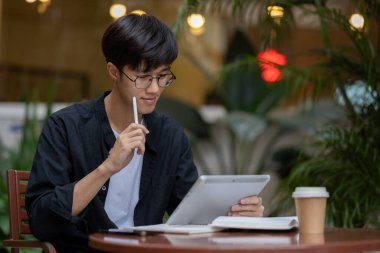 Yakışıklı, yakışıklı bir Asyalı, kahve dükkanındaki açık hava masasında dijital tableti üzerinde çalışıyor. Uzaktan çalışan, modern şehir hayatı, insanlar ve teknoloji kavramları