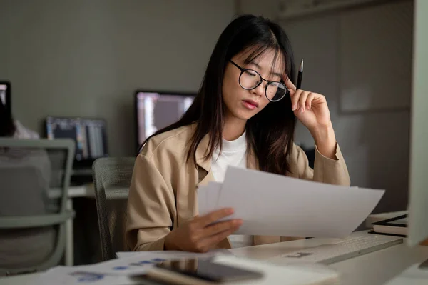 A stressed Asian businesswoman is reading a business report at her desk with a serious face, feeling tired and overworked.