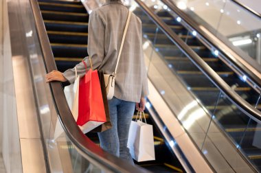 Alışveriş poşetleriyle modaya uygun giyinmiş bir kadının görüntüsü bir alışveriş merkezinde yürüyen merdivene çıkıyor. şehir hayatı, tüketim, yaşam tarzı, güzellik ve moda kavramları