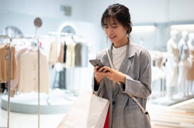 Moda kıyafetleri içinde mutlu, güzel bir Asyalı kadın hafta sonları alışveriş merkezinde yürürken telefonundaki mesajları kontrol ediyor. Yaşam tarzı, güzellik ve moda kavramları