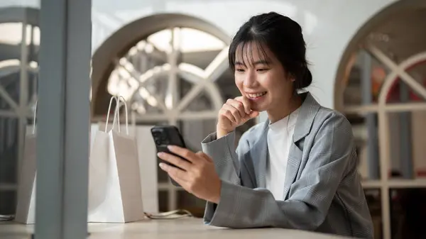 A charming Asian woman in trendy clothed is using her smartphone while relaxing in a cafe in the city. city life, wireless technology, and lifestyle concept