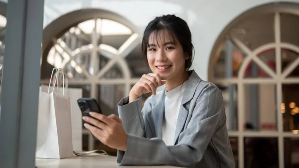 A charming Asian woman in trendy clothed is using her smartphone while relaxing in a cafe in the city. city life, wireless technology, and lifestyle concept