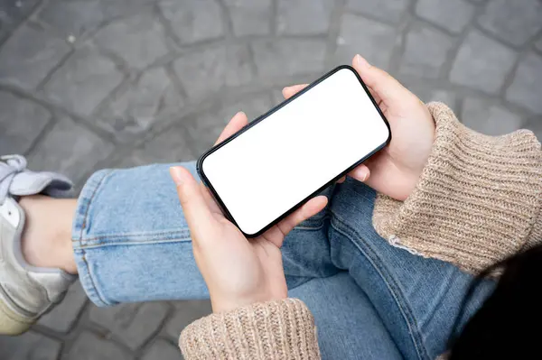 A woman is watching a video on her smartphone while sitting on a bench outdoors, holding a phone in the horizontal position. a white-screen smartphone mockup. people and technology concepts