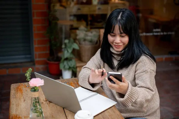 A positive Asian woman is chatting with her friends or checking messages on her phone while sitting at a table in a cafe in the city. People and modern city life concepts