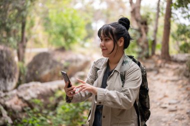 Sırt çantalı olumlu bir Asyalı kadın dağda yürüyüş yaparken akıllı telefonunu kullanıyor. İnsanlar, yaşam tarzı ve teknoloji kavramları