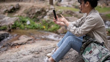 Pozitif Asyalı bir bayan gezginin yan görüntüsü bir kayanın üzerinde oturuyor ve akıllı telefonunu kullanıyor, dağda yürüyüş yaparken mola veriyor..
