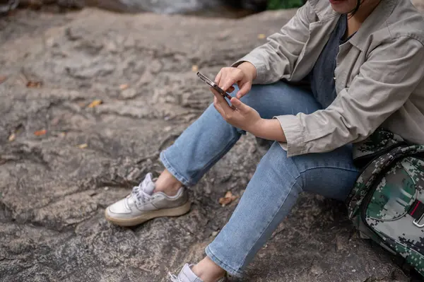 A cropped shot of an Asian woman using her phone, searching for information, or looking at the map on her phone while taking a break during her hiking trip.