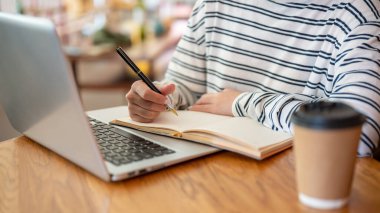 Bir kadının ödev yaparken, yazarken ya da kahve dükkanındaki bir masada not alırken yakın plan görüntüsü. Uzaktan çalışma, serbest çalışma, üniversite öğrencisi