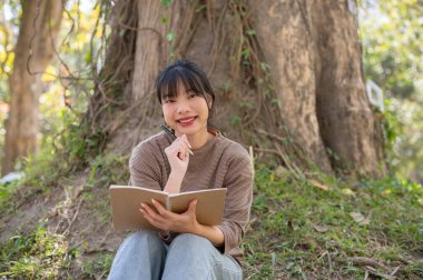 Çekici genç bir Asyalı kadın kitap okuyor ve parkta bir ağacın altında dinlenirken ilhamını buluyor. Boş zaman, yaşam tarzı.