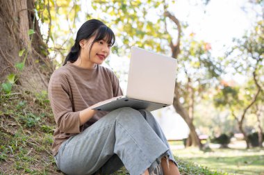 Güzel genç Asyalı bir kadın dizüstü bilgisayarını kullanıyor, parlak bir günde parktaki bir ağacın altında çalışıyor. İnsanlar ve kablosuz teknoloji kavramları