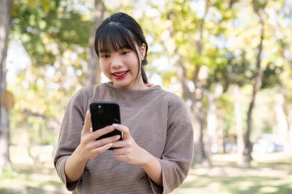 A beautiful young Asian woman is chatting with her friends on her smartphone while walking in the park on a bright day. reading messages, responding chat, on the website