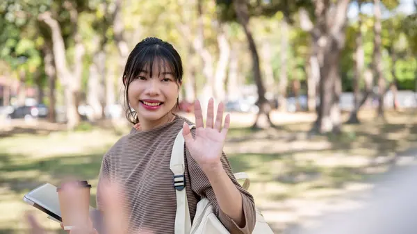 A friendly, cute young Asian female college student is waving her hand, greeting, or saying goodbye to her friend in a park. university life, school