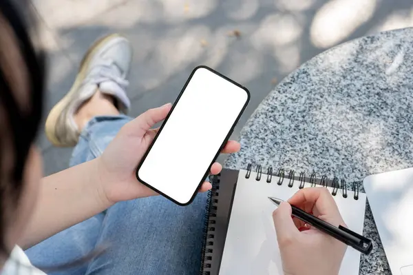 一个亚洲女人坐在户外餐桌前 用智能手机在笔记本上记笔记的特写镜头 白屏智能手机的模型 — 图库照片