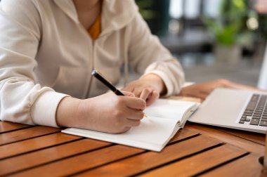 Yakın plan bir resim, beyaz kapüşonlu bir kadının açık hava kafede çalışırken kitaptaki fikirlerini yazdığını gösteriyor..