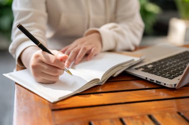 Kesilmiş bir resimde, açık hava kafesinde uzaktan çalışan beyaz kapüşonlu bir kadın, fikirlerini bir kitapta sıralıyor ve dizüstü bilgisayarını kullanıyor..