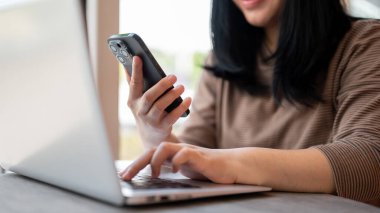 Akıllı telefonu ve dizüstü bilgisayarını kullanan Asyalı bir kadının yakın plan fotoğrafı. Bir kafede ortak çalışma alanında çalışıyor. İnsanlar ve kablosuz teknoloji kavramları