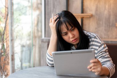 Stresli bir Asyalı kadın ciddi bir yüz ifadesiyle tablet ekranına bakar ve bir kafede uzaktan çalışırken sorunu çözer..
