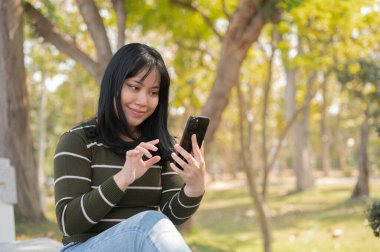 Yeşil bir parktaki bankta dinlenirken akıllı telefonunu kullanan genç bir Asyalı kadın. İnsanlar ve kablosuz teknoloji kavramları